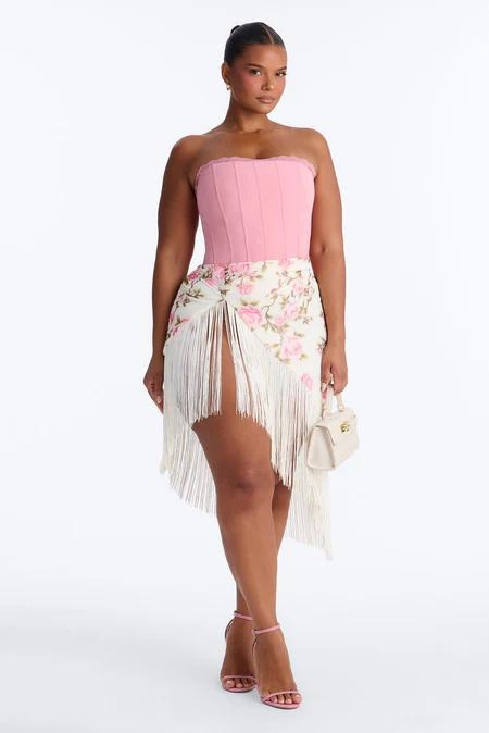 Mujer con top corsé rosa sin tirantes, falda blanca asimétrica con estampado floral y flecos, tacones rosas, bolso beige.