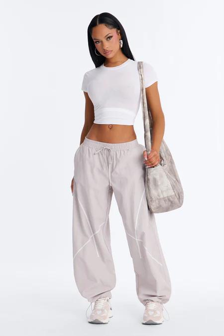 Woman wearing a white crop top, light grey baggy track pants with white stitching, and light sneakers, holding a grey shoulder bag. Full shot.