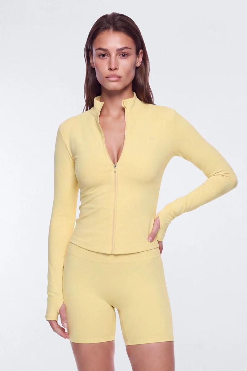 Woman wearing a pastel yellow activewear set with a long-sleeve zip-up jacket and matching biker shorts from the new athletic collection.