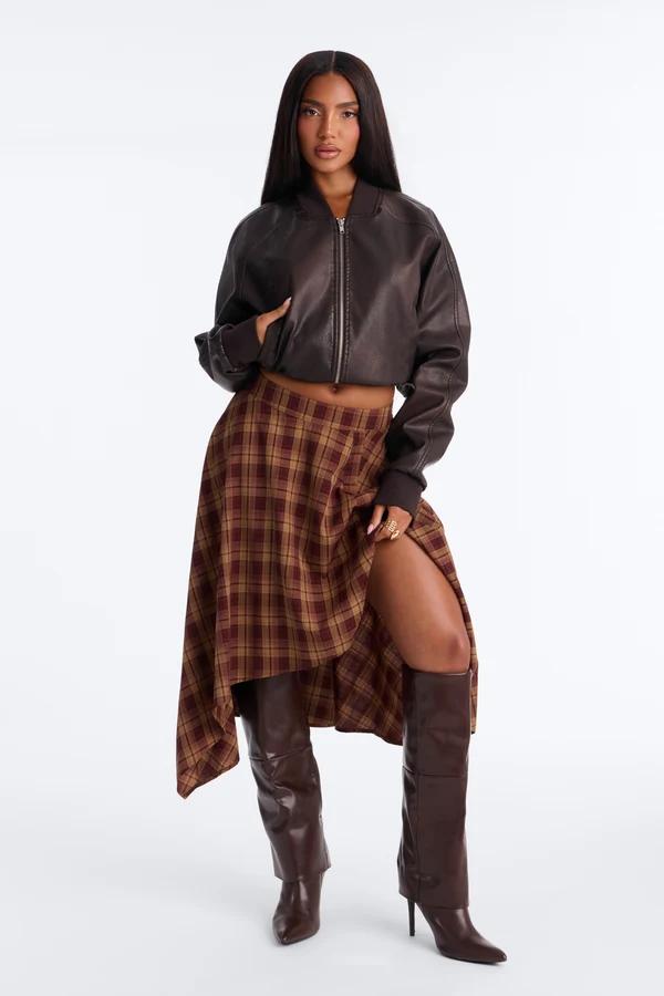 Woman in a brown leather bomber jacket, plaid midi skirt, and high boots, promoting a chic autumn fashion collection on a white background.