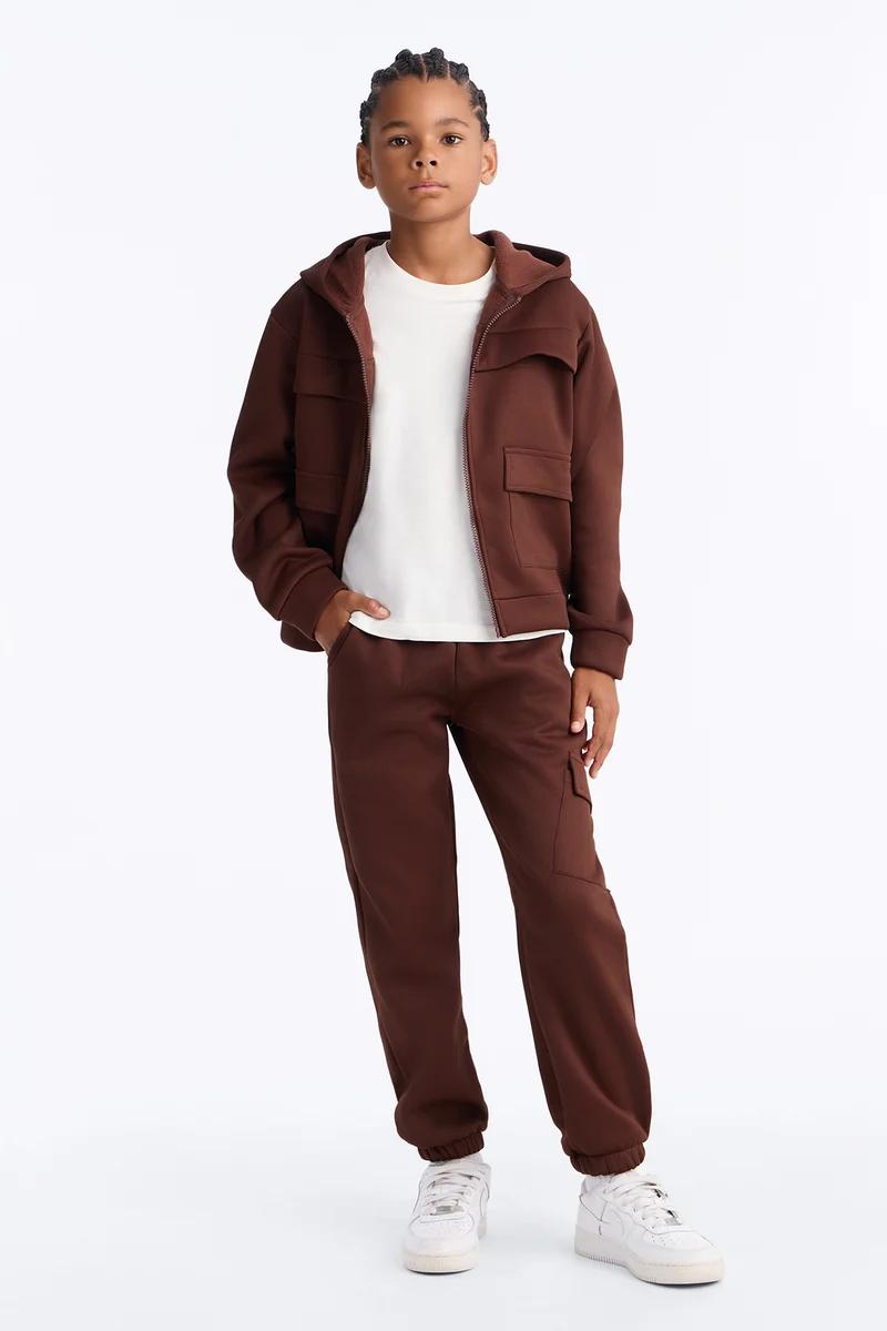 Niño con chándal marrón infantil de sudadera con capucha con cremallera, pantalones jogger cargo y tenis blancos.