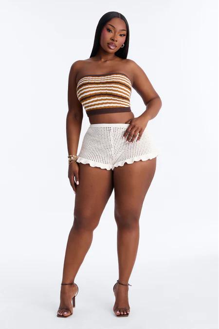 Mujer en top corto de crochet a rayas (marrón, dorado, blanco) y shorts de crochet blancos con volantes. Lleva tacones marrones y pulsera dorada.