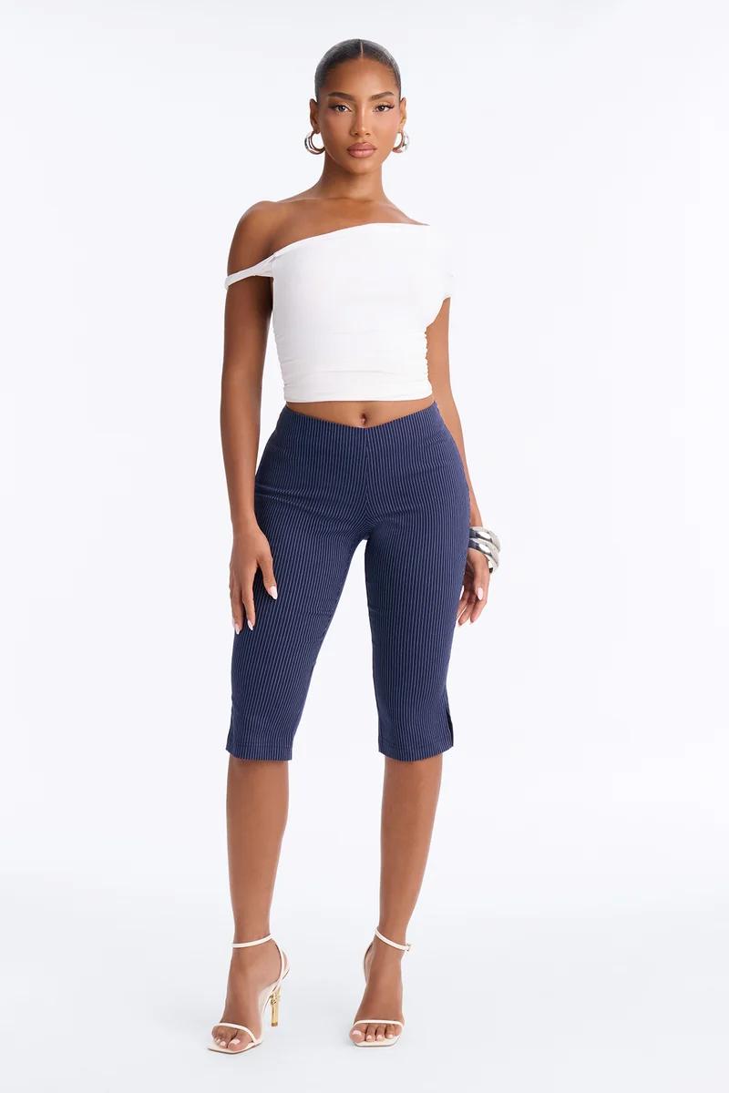 Woman posing in a white off-the-shoulder crop top, navy blue pinstripe capri pants, and white high heels.