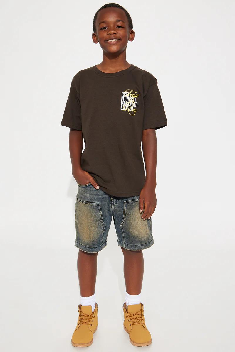 Smiling Black boy in brown graphic tee "PRAY TONIGHT & LEAVE IT ALONE", distressed denim shorts, and tan boots.