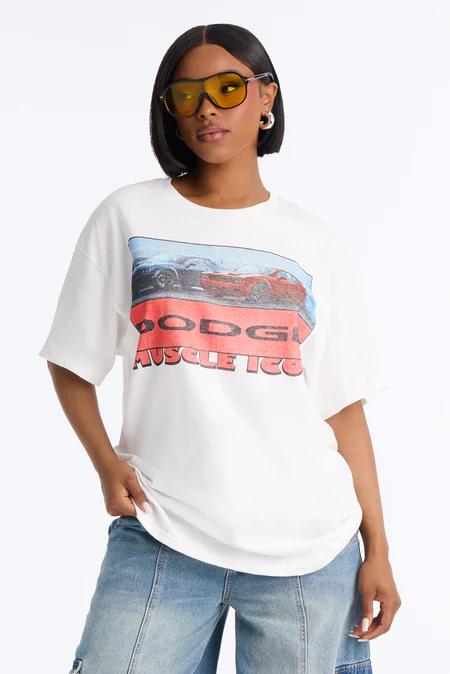 Woman in yellow sunglasses, white oversized t-shirt with a "DODGE MUSCLE 100" graphic & two sports cars, and light wash jeans.