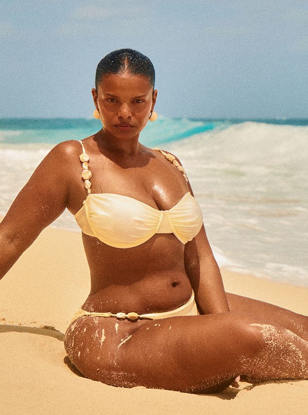 Woman wearing a yellow strapless bikini with shell accents, sitting on a sandy beach. Perfect for stylish summer swimwear.