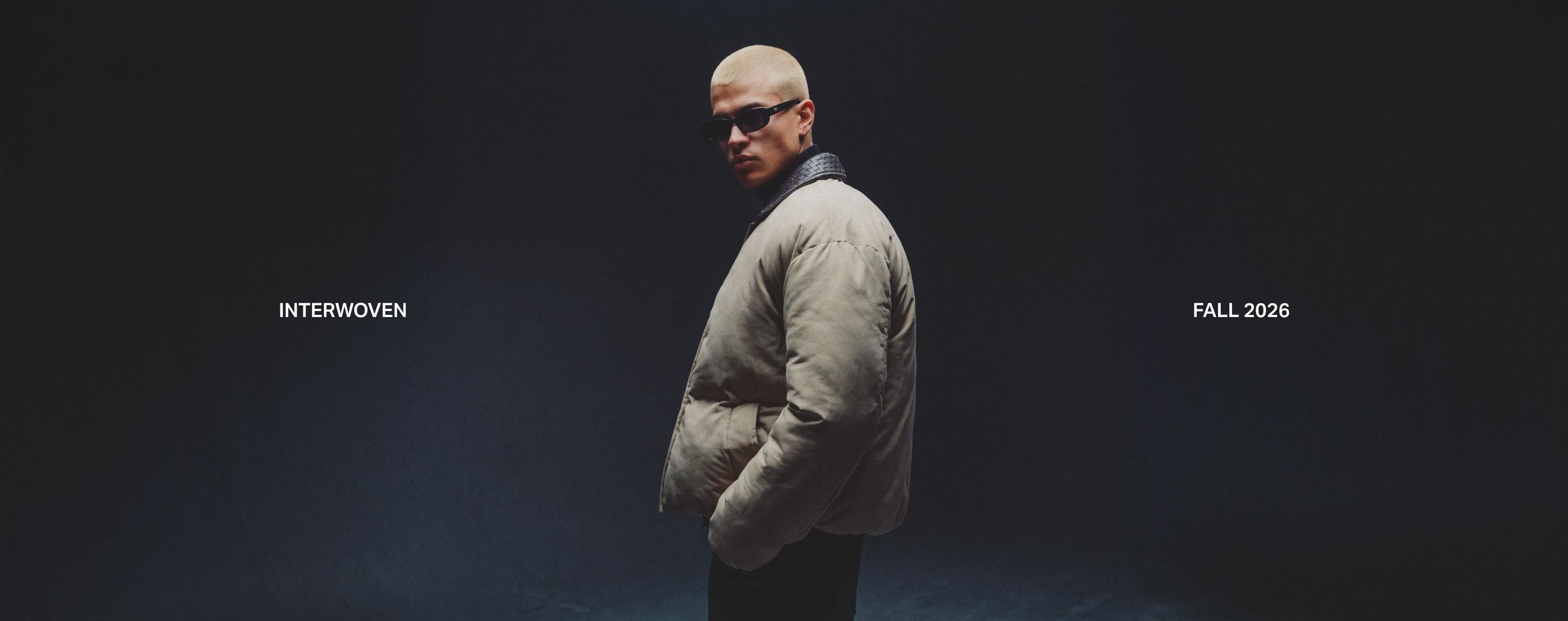 Model wearing black sunglasses and beige puffer jacket on dark studio backdrop promoting Interwoven Fall 2026 outerwear collection.