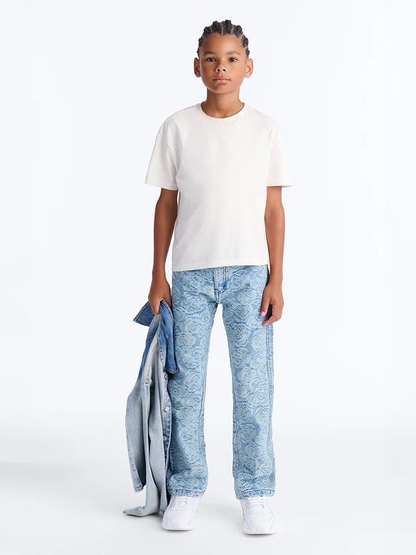 Young boy models a white t-shirt and light blue floral jeans, holding a denim jacket. Shop our trendy kids' casual wear.