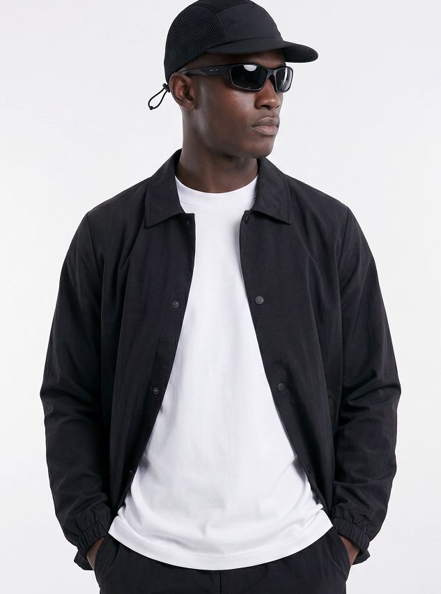 Hombre con estilo modela chaqueta coach negra casual, camiseta blanca, gorra y gafas de sol, luciendo moda urbana moderna.