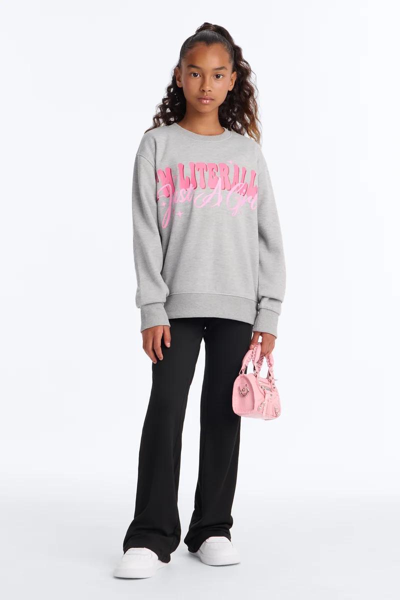 Young girl wearing a gray graphic sweatshirt, black flared pants, and holding a small pink handbag.