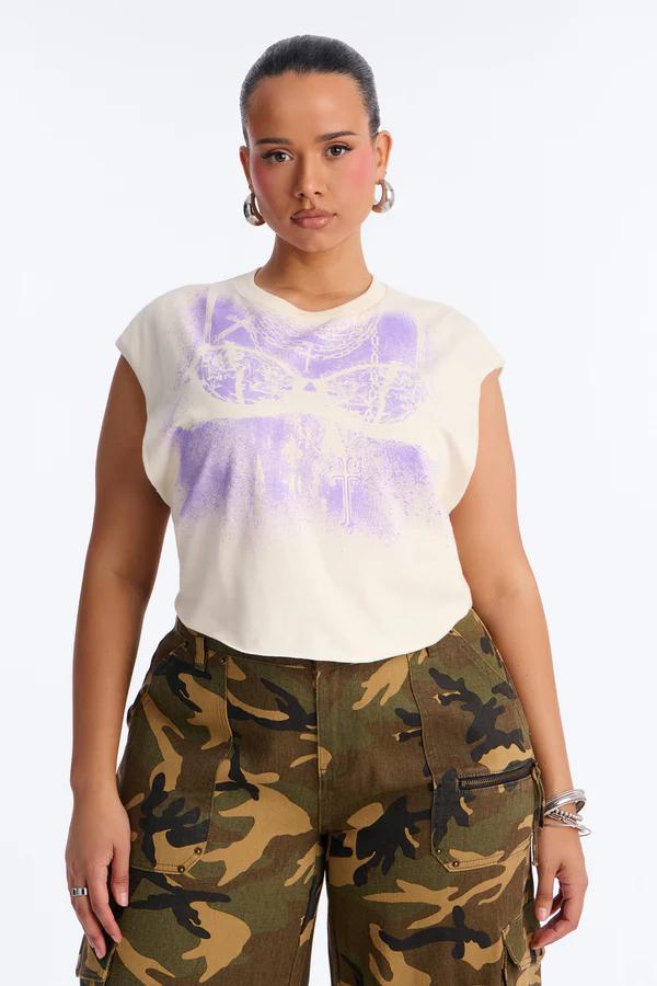 Persona con camiseta crema de manga corta con gráfico púrpura desgastado de sujetador de cadenas y ankh, con pantalones cargo de camuflaje verde.