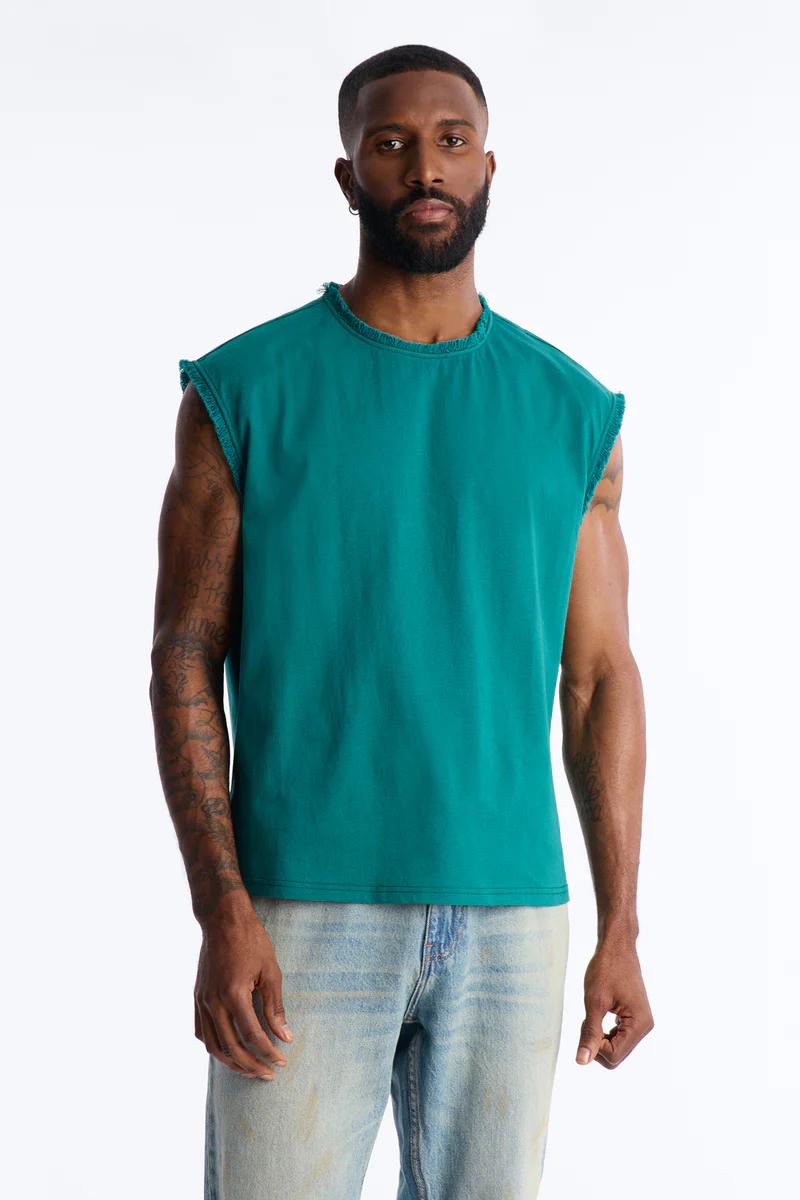 Man models a vibrant teal sleeveless t-shirt with distressed edges and light wash jeans, perfect for trendy men's summer casual wear.
