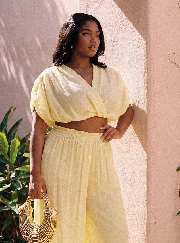 Curvy woman in light yellow puff-sleeve crop top & wide-leg pants, holding a gold structured handbag. Chic summer resort wear fashion.