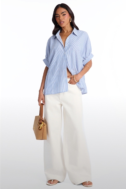 Mujer con camisa de rayas azules oversize, pantalones blancos anchos y bolso camel, mostrando un look casual chic de verano.
