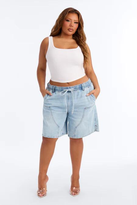 Mujer con top corto blanco cuello cuadrado, shorts denim azul claro con cordón y cintura elástica, y mules transparentes.