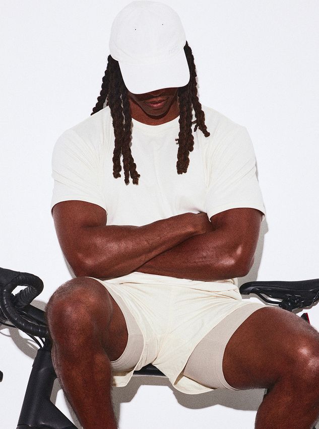 Persona con gorra blanca y ropa activa crema, rastas, sentada en bicicleta estática, brazos cruzados. Presenta ropa deportiva masculina versátil.