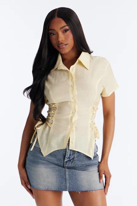 Woman in a sheer pale yellow short-sleeve button-up shirt with side corset lacing, paired with a blue denim mini skirt, looking forward.
