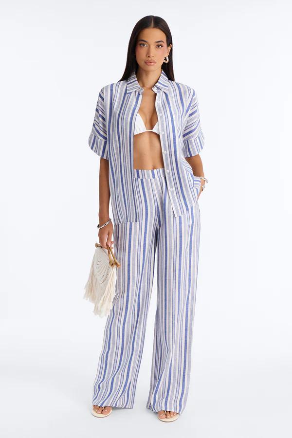 Woman modeling a cream cropped shirt, wide-leg pants, and a pink/yellow floral sarong with white lace. She wears sunglasses and gold heels.