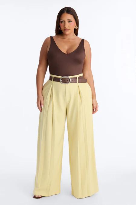 Mujer con top marrón de cuello en V, pantalones anchos plisados amarillos claros de cintura alta y cinturón marrón con hebilla plateada.