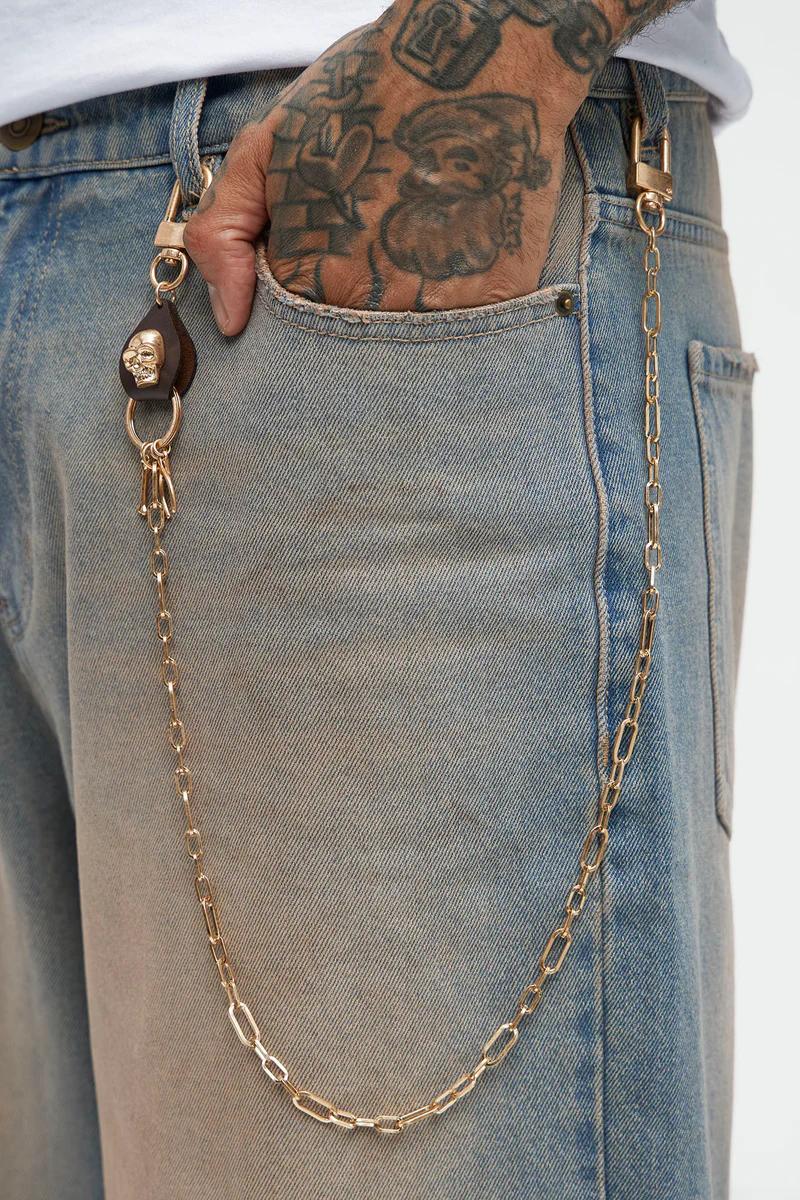 Tattooed hand in faded blue jeans pocket, golden wallet chain with brown leather skull keychain. Showcasing stylish accessories.