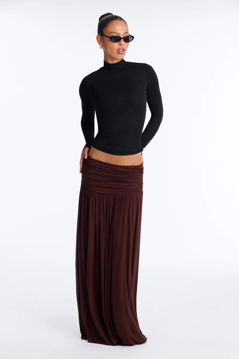 Model in a black long-sleeve turtleneck top and low-rise brown ruched maxi skirt with sunglasses.