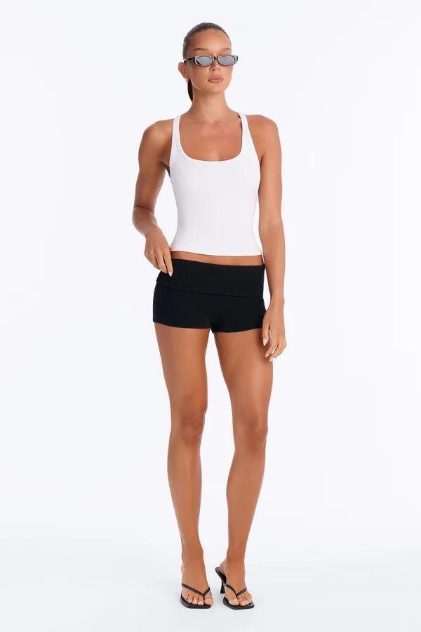 Model in a white ribbed tank top and black fold-over shorts showcases the new summer activewear collection against a white background.