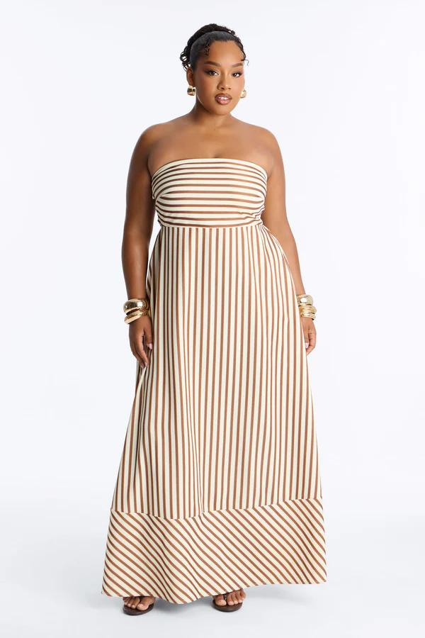 Smiling woman models an elegant brown/beige striped strapless maxi dress with diagonal hem, ideal plus-size summer fashion.