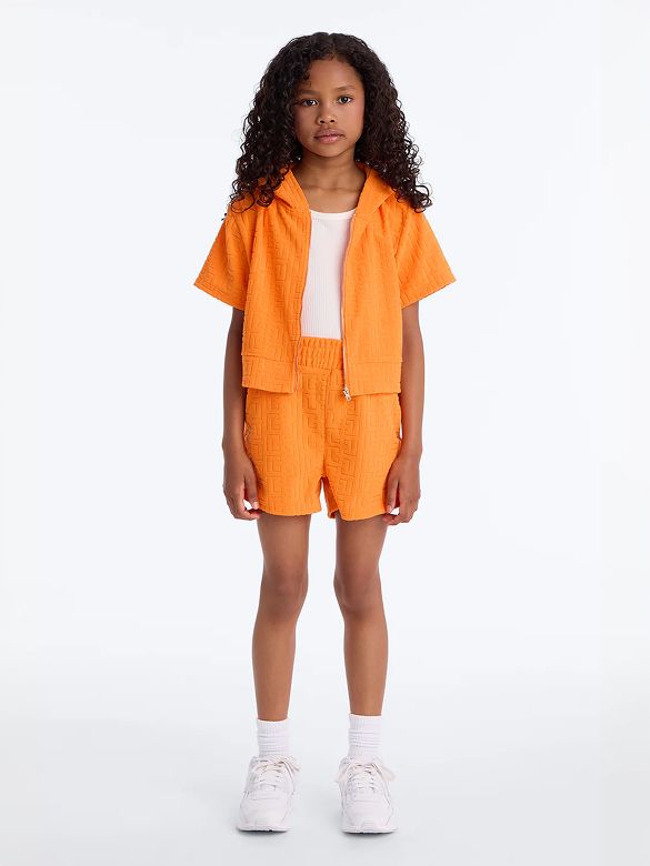 Young girl in an orange patterned terry cloth hoodie and matching shorts, white tank, and sneakers. Explore comfy kids' summer fashion.