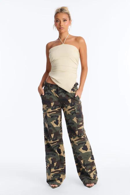 Full-length shot: woman in beige halter crop top & loose camouflage cargo pants, hands in pockets, white background.