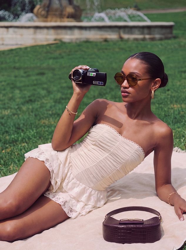 Woman in a strapless cream summer dress and retro sunglasses holding a vintage camera on a picnic blanket, showcasing a new fashion collection.