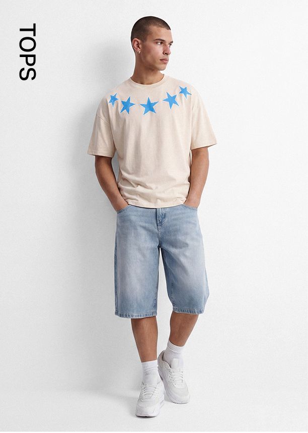 Hombre en camiseta beige extragrande con estrellas azules, shorts de mezclilla claros y zapatillas blancas, mostrando tops masculinos.