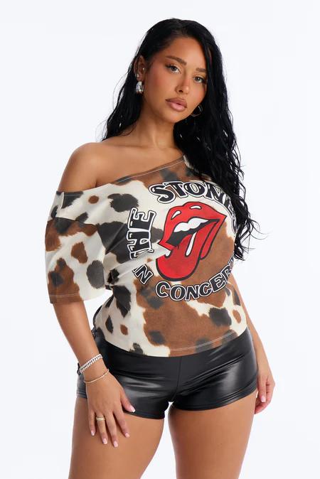 Woman wearing an off-shoulder cow print top with Rolling Stones logo, black leather shorts, silver jewelry against white.