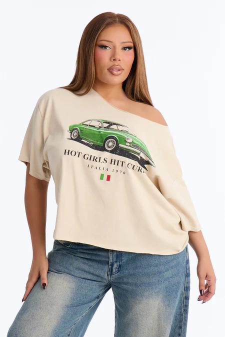 Woman posing in a beige off-shoulder tee featuring a green vintage car graphic and "HOT GIRLS HIT CURVES" text, with blue jeans.