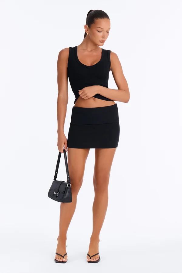 Woman posing in a black ribbed tank top and low-rise miniskirt with a matching handbag, showcasing trendy monochrome summer fashion styles.