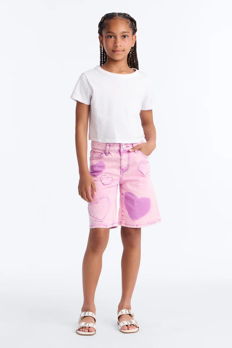 Girl in white crop top, pink acid wash heart denim shorts, white sandals. Kids summer fashion.