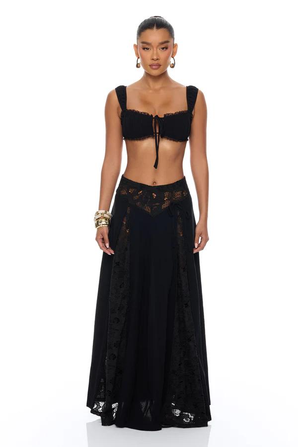 Woman standing in a bronze open-knit crochet set: ruffled long-sleeve top, flared pants, sunglasses, and clutch.