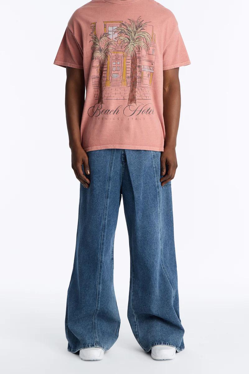 Hombre con camiseta gráfica rosa palo "Beach Hotel Venice", jeans anchos. Promoción de moda casual de verano para hombre.
