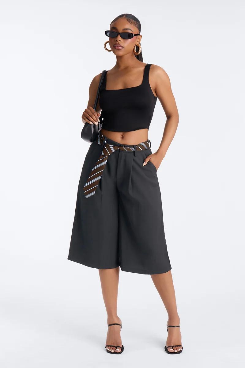 Woman in a black crop top, dark wide-leg bermuda shorts with a striped tie belt, sunglasses, and heels.