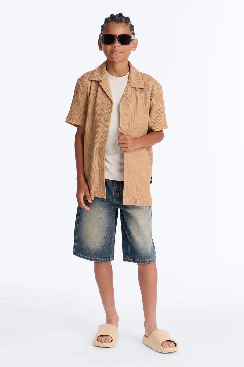 Niño modela atuendo casual de verano: camisa beige, shorts de mezclilla desgastados, sandalias beige, gafas.