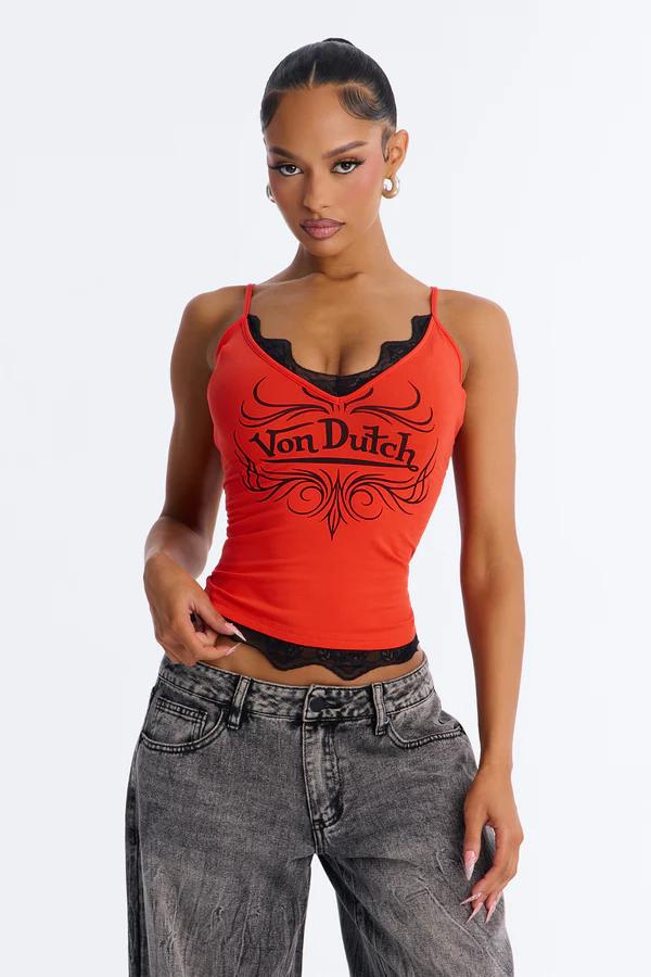 Woman in a red Von Dutch spaghetti strap top with black lace, paired with grey acid-wash wide-leg jeans, posing against a white background.