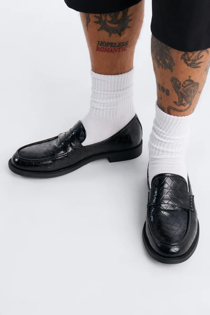 Stylish black textured men's loafers paired with white socks, showcasing modern fashion footwear on a clean white background.