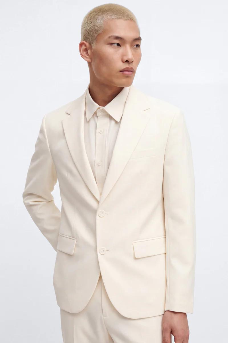 Man wearing a stylish cream two-button suit jacket and matching trousers, with a pale cream shirt, showcasing modern men's formalwear.