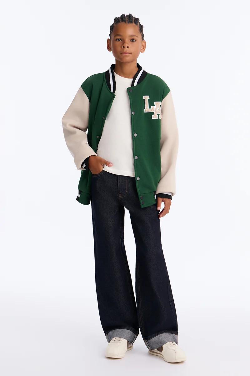 Boy wearing a green and cream kids varsity jacket with LA patch, white t-shirt, and dark wide-leg jeans.