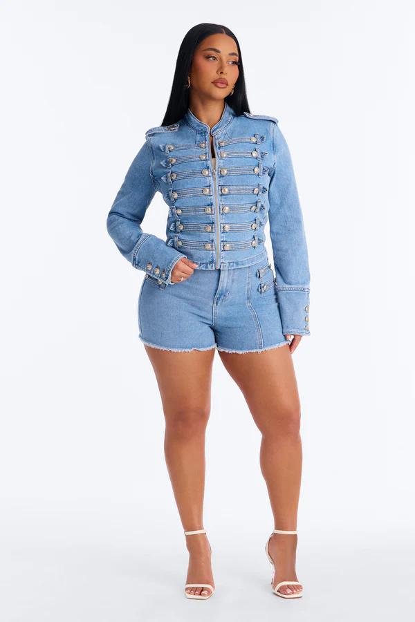 Woman in a light blue denim military-style jacket with silver buttons and matching frayed shorts, standing full-body against a white background.