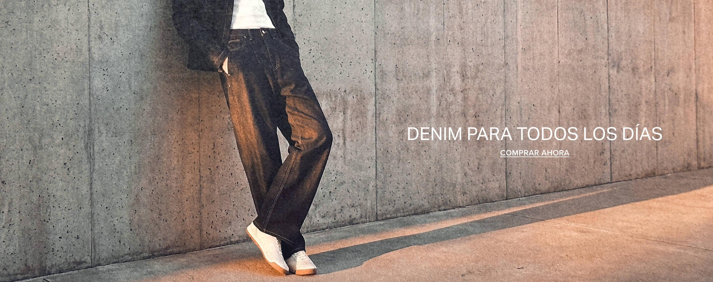 Persona con vaqueros anchos oscuros y tenis blancos apoyada contra una pared de concreto. Texto superpuesto: Everyday Denim, Shop Now.