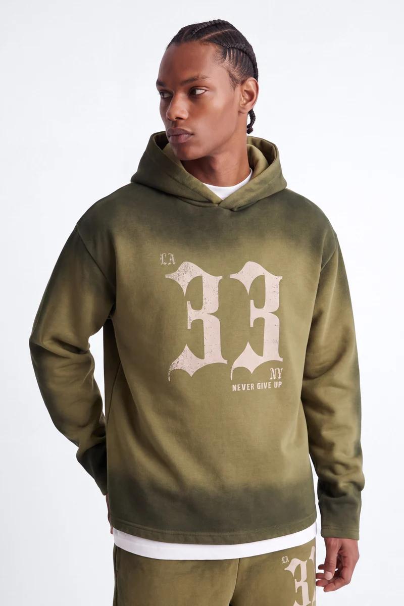 Man wearing oversized olive green graphic hoodie with "LA 33 NY NEVER GIVE UP" design, fading to darker green. Ideal men's streetwear.