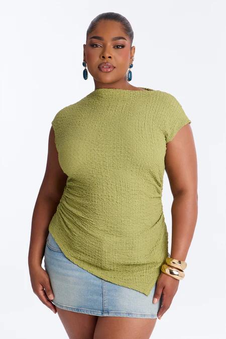 Mujer con blusa texturizada verde oliva asimétrica de manga corta, minifalda de mezclilla clara, aretes azules y pulseras doradas.