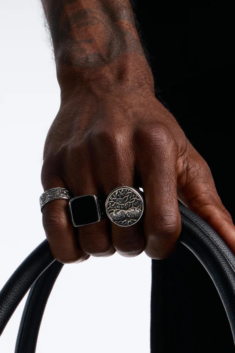 Mano mostrando tres llamativos anillos para hombre: banda grabada, sello rectangular negro y anillo de plata del Árbol de la Vida. Ideal para colección de joyas.