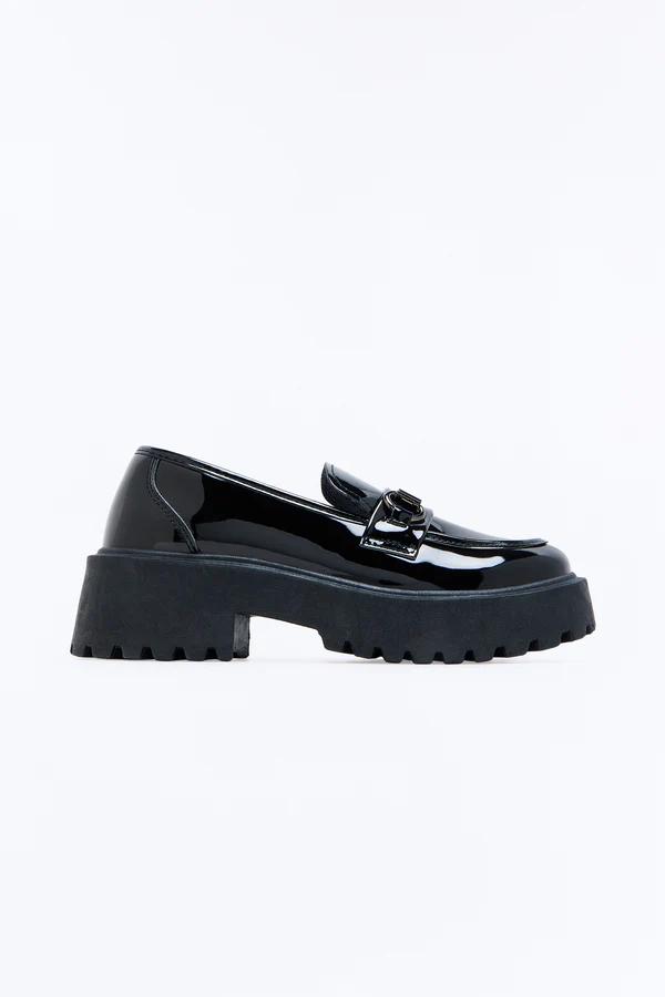 Mocasín elegante de charol negro con una hebilla plateada y una suela gruesa y texturizada, sobre un fondo blanco limpio.