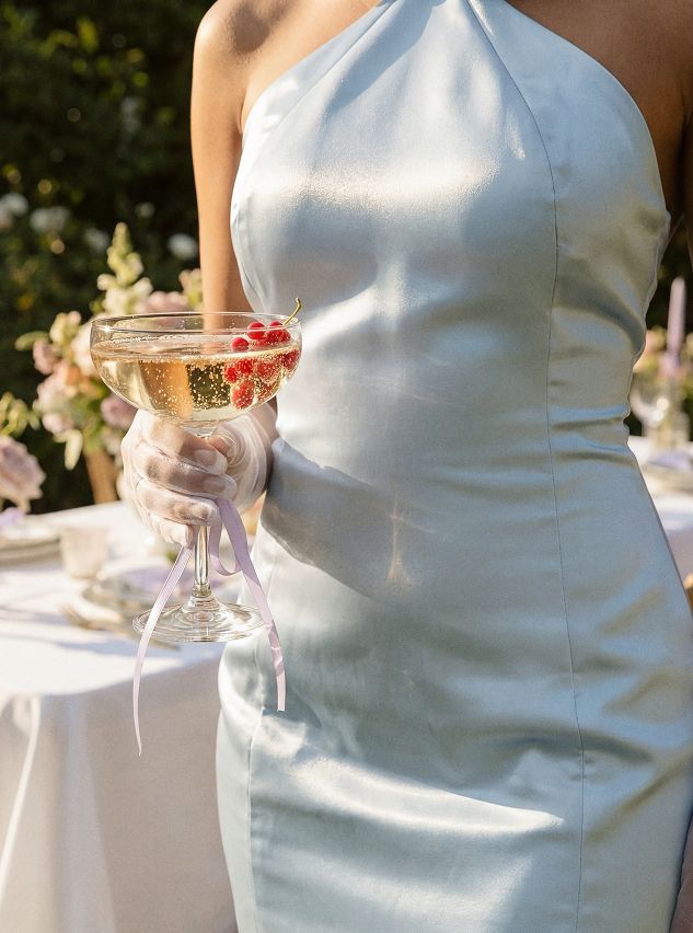 Modelo elegante con vestido de satén azul claro y guantes sostiene copa de champán con grosellas rojas, promocionando vestimenta de evento refinada.